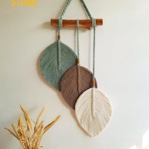 Pode incluir: Um pendurador de parede em macramé com três desenhos em forma de folha em tons de verde, castanho e creme. As folhas são suspensas por uma barra de madeira e um cordão verde trançado. O texto "TURN STUDIO" é visível no canto superior esquerdo.