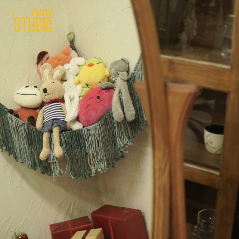 May include: A collection of plush toys, including a red and black frog, a striped bunny, a white bunny, a yellow chick, and a gray cat, are arranged in a green fringed hammock. The hammock is hanging on a white wall. The text "Tuny Studio" is visible in the top left corner of the image.