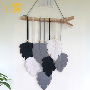 May include: A macrame wall hanging featuring seven leaf-shaped designs in black, white, and gray. The leaves are suspended from a wooden branch with wooden beads and hanging cords. The text "TINY STUDIO" is in the upper left corner.