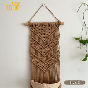 May include: A handcrafted macrame wall hanging in a natural tan color. The design features a geometric pattern with a wooden dowel at the top for hanging. The bottom has a small pocket. The text "TUNY STUDIO" is in the upper left corner, and "Style 3" is in the lower right.
