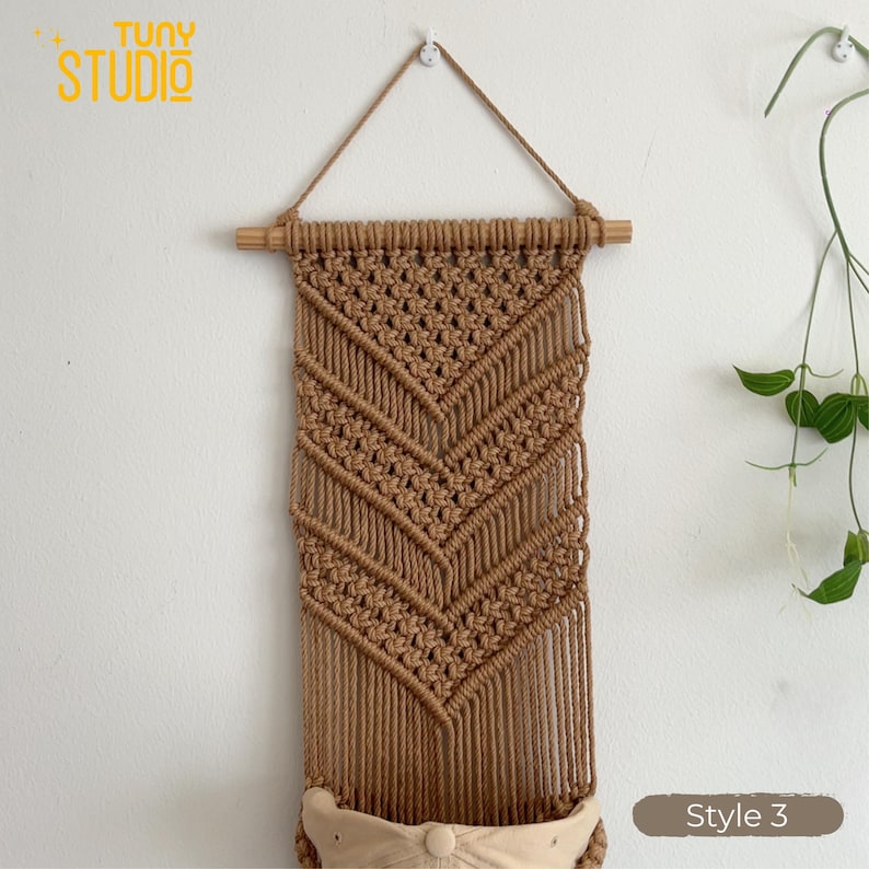 May include: A handcrafted macrame wall hanging in a natural tan color. The design features a geometric pattern with a wooden dowel at the top for hanging. The bottom has a small pocket. The text "TUNY STUDIO" is in the upper left corner, and "Style 3" is in the lower right.