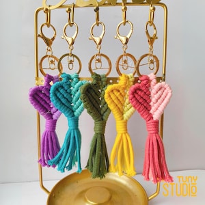 May include: A collection of macrame heart-shaped keychains in various colors, including purple, teal, green, yellow, and pink. Each keychain has a gold-colored clasp and a tassel. The keychains are displayed on a gold-colored stand. The text "TUNY STUDIO" is visible.