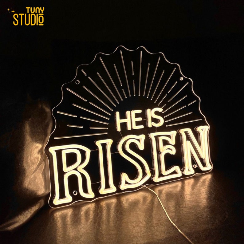 May include: A black neon sign with a sunburst design and the words "He is Risen" in white neon.