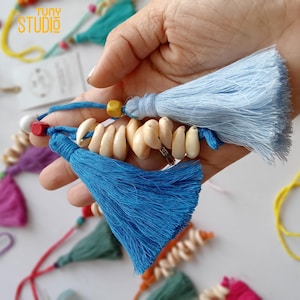 May include: Two keychains with blue and light blue tassels, white seashells, and yellow and red beads. The keychains are held in a hand.