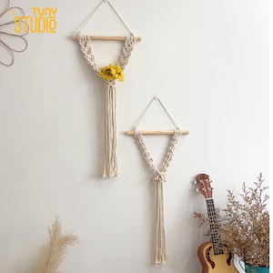 May include: Two macrame wall hangings with wooden dowels and cream-colored rope. One features a yellow sunflower. The hangings are triangular in shape and suspended from the wall. The logo "TUNY STUDIO" is in the upper left corner.