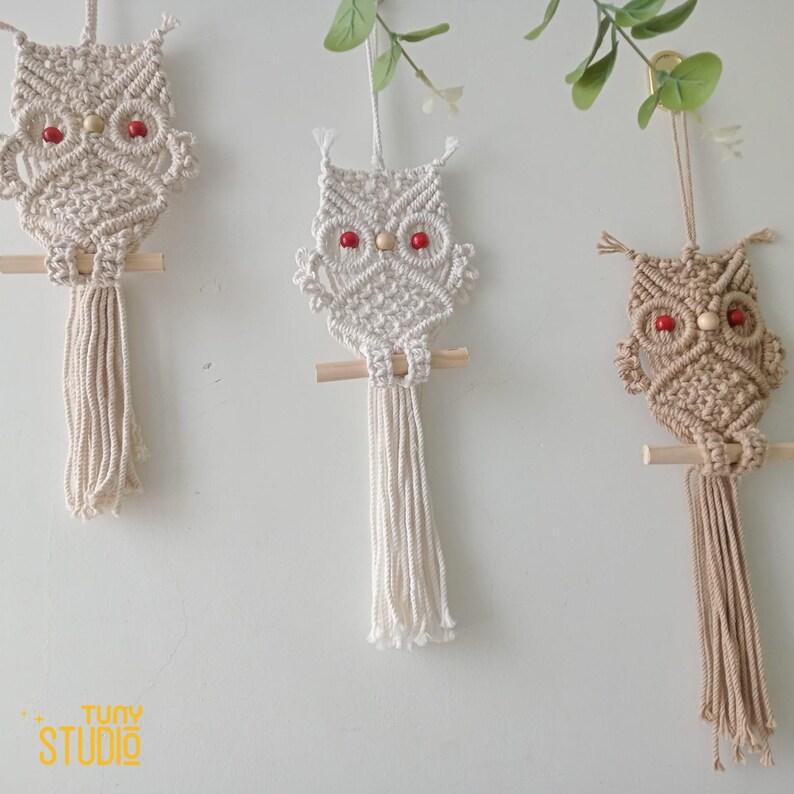 K&ouml;nnte beinhalten: Drei Makramee-Eulen-Wandbeh&auml;nge mit Holzst&auml;ben und roten Perlenaugen. Die Eulen sind in wei&szlig; und beige gehalten.