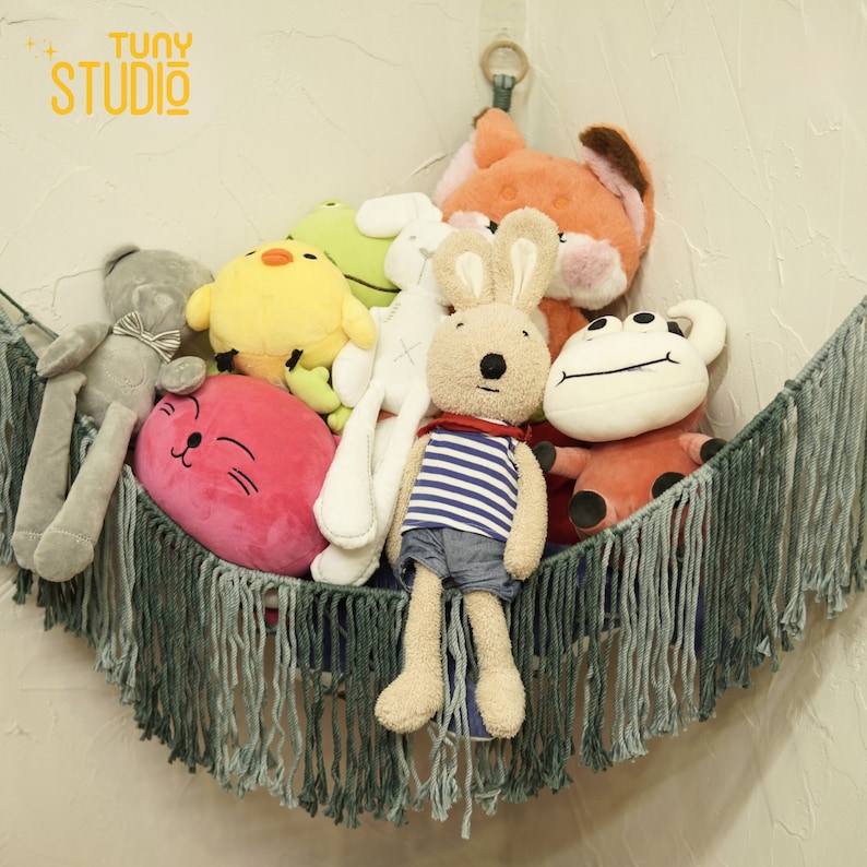 May include: A collection of plush toys in a green fringe hammock. The toys include a grey bunny, a pink cat, a yellow chick, a green frog, a white bunny, an orange fox, and a white and red frog.