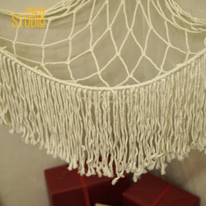 May include: A white macrame hammock with a fringe border. The hammock is made of a thick, knotted rope and has a natural, bohemian style.