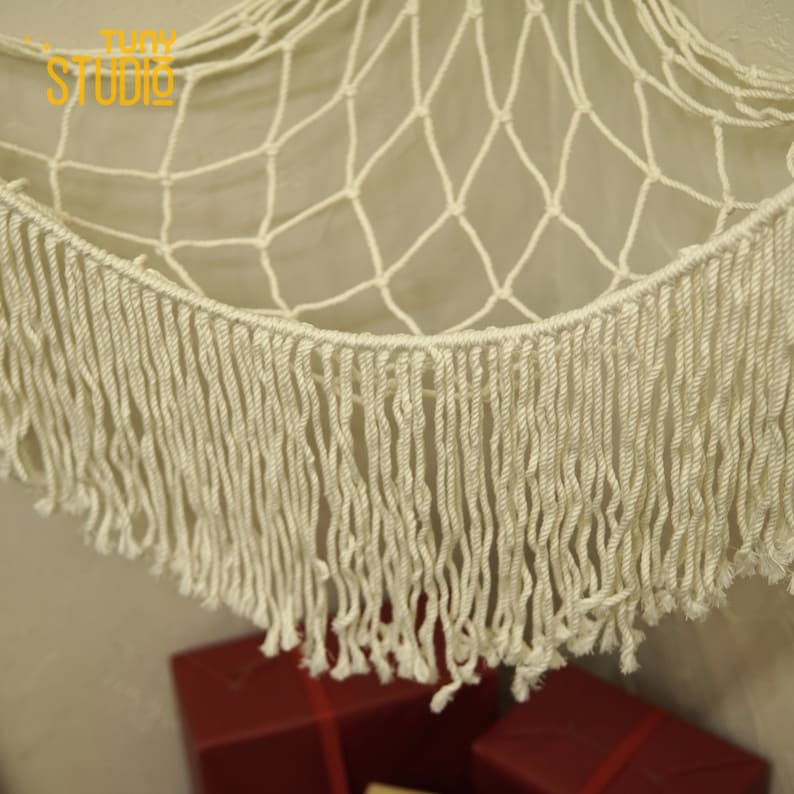May include: A white macrame hammock with a fringe border. The hammock is made of a thick, knotted rope and has a natural, bohemian style.
