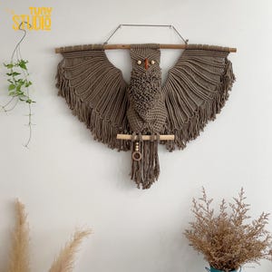 Puede incluir: Un colgante de pared de macramé con forma de búho en tonos marrones, con alas extendidas y una percha de madera. El búho presenta un trabajo de nudos detallado, cuentas de madera y detalles en naranja para los ojos. El texto "TUNY STUDIO" es visible.