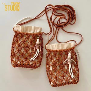 May include: Two small, handmade macrame bags in a warm brown colour. Each bag has a cream-coloured lining and a decorative wooden doll charm. The bags have long, braided brown straps for wearing over the shoulder. The text "TUNY STUDIO" is visible in the top left corner.