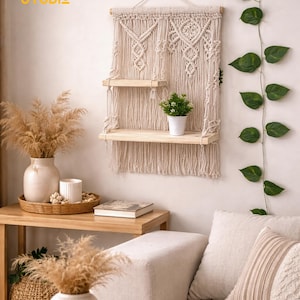 Puede incluir: Una estantería de pared de macramé con dos estantes de madera, una pequeña planta en maceta y un diseño tejido beige. La estantería de pared está colgada en una pared blanca. El texto "TUNY STUDIO" está en la esquina superior izquierda.