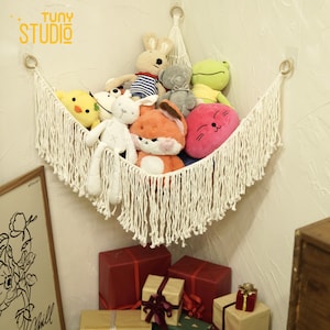 May include: A white macrame hammock with fringe hanging from a corner wall, filled with stuffed animals including a yellow chick, a white bunny, a red fox, a gray bear, a green frog, and a pink cat with a smiling face.