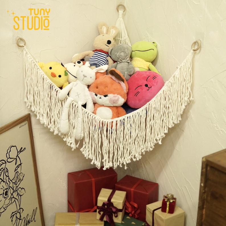 May include: A white macrame hammock with fringe hanging from a corner wall, filled with stuffed animals including a yellow chick, a white bunny, a red fox, a gray bear, a green frog, and a pink cat with a smiling face.