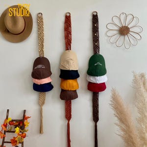 May include: A set of three macrame wall organizers with wooden rings, each holding a stack of baseball caps in different colors. The caps are brown, navy blue, pink, white, green, red, and yellow.