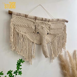 May include: A macrame wall hanging in the shape of an elephant, with a wooden dowel at the top and a fringe around the edges. The elephant is made of natural colored cotton cord.