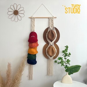 Peut inclure: Porte-chapeaux en macramé avec une tringle en bois et une corde de suspension. Il présente une pile de casquettes de baseball colorées et deux chapeaux fedora bruns. Une décoration murale en forme de marguerite et un vase avec de la verdure sont également visibles.