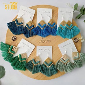 Puede incluir: Seis pares de pendientes con flecos de borla. Los pendientes están hechos de un material marrón claro con un diseño geométrico. Las borlas son en tonos de azul y verde.