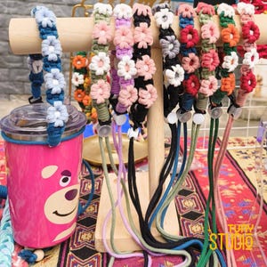 May include: A display of colorful handmade lanyards with floral designs. The lanyards are in various colors, including blue, pink, white, and black. A pink tumbler with a bear graphic is on the left. The lanyards are displayed on a wooden stand.