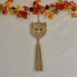 Może przedstawiać: Beżowy makramowy wiszący na ścianie sowa z długim frędzlem. Sowa ma szczegółowy tkany wzór z dwoma czarnymi oczami z koralików i drewnianym noskiem z koralików. Widoczny jest napis "STUDIO".