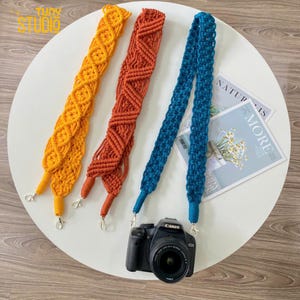 Puede incluir: Tres correas de cámara hechas de macramé tejido en amarillo, naranja y azul. Las correas están colgadas en una superficie blanca con una cámara en primer plano.