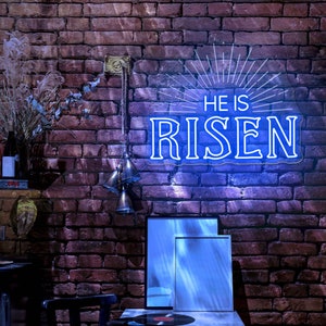 May include: A neon sign with a blue glow that reads "He is Risen" with a sunburst design behind the text. The sign is hanging on a brick wall with a wooden shelf and a few decorative bells hanging below the sign.