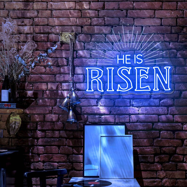 May include: A neon sign with a blue glow that reads "He is Risen" with a sunburst design behind the text. The sign is hanging on a brick wall with a wooden shelf and a few decorative bells hanging below the sign.