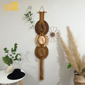 May include: A macramé hat holder displaying three fedora hats in brown and beige tones. The hats are suspended from a wooden bar, featuring woven details and a long fringe. The wall decor includes a metal flower and greenery.