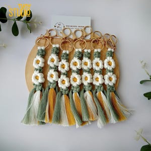 May include: Set of seven green, white and yellow macrame keychains with daisy flower designs and gold key rings. The keychains are displayed on a wooden surface. "WOOD AND ROPES - MACRAME" is written on a white card.