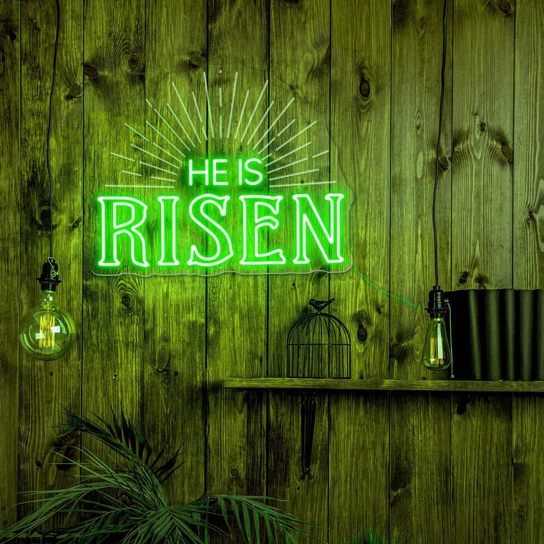 May include: Green neon sign with the words "He is Risen" on a wooden wall with two light bulbs hanging from the ceiling and a birdcage on a shelf.