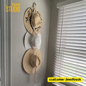 May include: Three straw hats of different colours and styles are hanging on a wall using a macrame rope hanger. The hats are stacked on top of each other. The wall is white and the background is a white window blind.