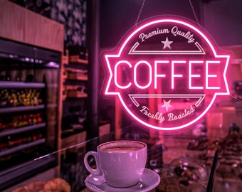 May include: A pink neon sign with the words "Premium Quality" and "Freshly Roasted" surrounding the word "COFFEE" in a larger font. A white coffee cup sits on a glass table in front of the sign.