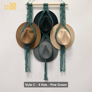 May include: A wall-mounted hat rack with four hats hanging from it. The rack is made of macrame with a pine green color. The hats are brown, black, beige, and gray.