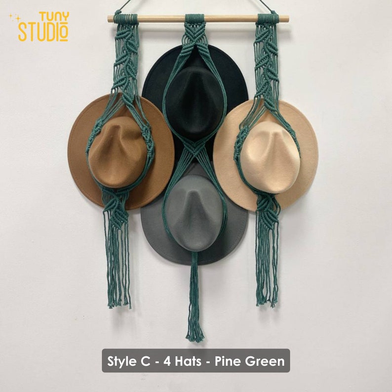 May include: A wall-mounted hat rack with four hats hanging from it. The rack is made of macrame with a pine green color. The hats are brown, black, beige, and gray.