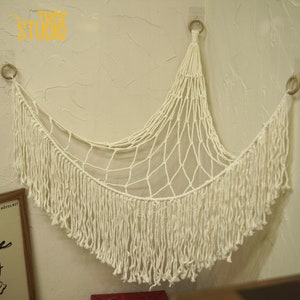 May include: White macrame wall hanging with a fringe border. The hanging is in the shape of a crescent moon and is made of cotton cord.