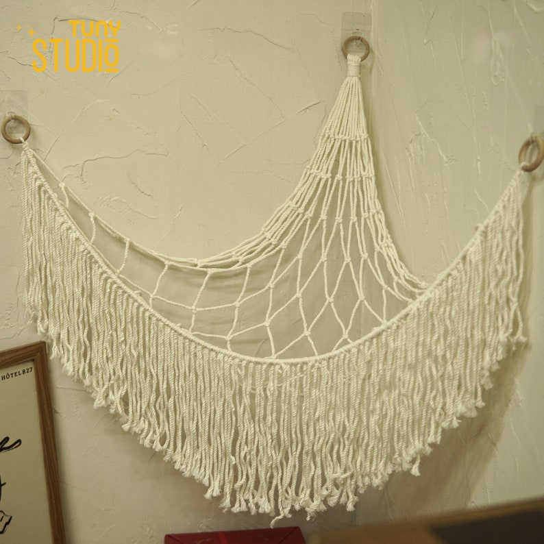 May include: White macrame wall hanging with a fringe border. The hanging is in the shape of a crescent moon and is made of cotton cord.