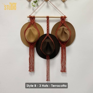 May include: Terracotta macrame hat holder with three hats hanging from it. The hat holder is made of a thick cord and has a wooden dowel at the top. The hats are brown, tan, and dark brown. The text "Style B - 3 Hats - Terracotta" is at the bottom of the image.