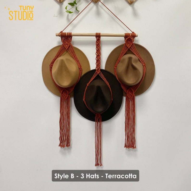 May include: Terracotta macrame hat holder with three hats hanging from it. The hat holder is made of a thick cord and has a wooden dowel at the top. The hats are brown, tan, and dark brown. The text "Style B - 3 Hats - Terracotta" is at the bottom of the image.