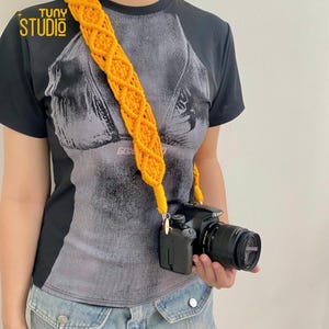 May include: A yellow crocheted camera strap with a black camera attached. The strap is worn over a person's shoulder.