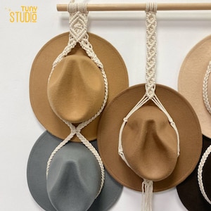 May include: A set of four hats hanging on a wooden dowel rod using macrame cord. The hats are brown, gray, and beige. The macrame cord is white.