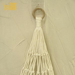 May include: A white macrame hanging with a wooden ring at the top. The macrame is hanging from a wall.