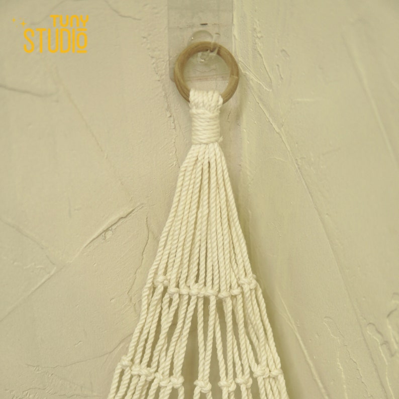 May include: A white macrame hanging with a wooden ring at the top. The macrame is hanging from a wall.