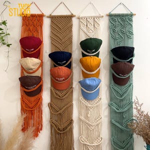 May include: Macrame wall hangings in orange, tan, cream, and green, each with multiple baseball caps in various colors. The caps have text that reads "NEW YORK" and "PARIS". The wall hangings are made of woven rope and are designed to hold the caps.