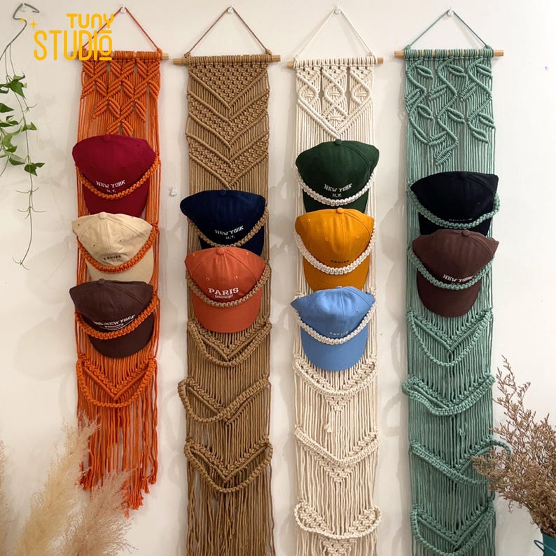 May include: Macrame wall hangings in orange, tan, cream, and green, each with multiple baseball caps in various colors. The caps have text that reads "NEW YORK" and "PARIS". The wall hangings are made of woven rope and are designed to hold the caps.