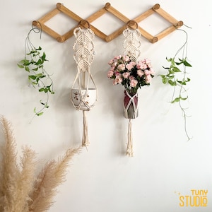 May include: A wooden expandable wall-mounted rack with two macrame plant hangers. One hanger holds a white ceramic pot with a floral design, and the other holds a vase with pink flowers. Green vines and pampas grass add to the decor.