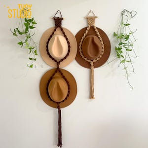 May include: Three fedora hats in beige, brown, and tan, displayed on a white wall. Each hat is suspended by a macrame rope hanger. The hats have a classic teardrop crown and a wide brim. The macrame hangers have a decorative tassel.