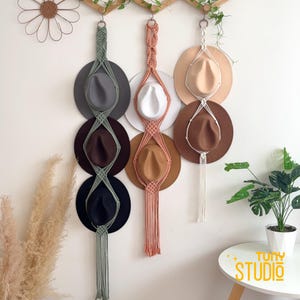 May include: Macrame hat holders in sage green, peach, and white, each holding two hats in various colours. The hats include grey, brown, black, white, tan, and dark brown. The holders are suspended from a wooden rod on a white wall, with a decorative flower and greenery.
