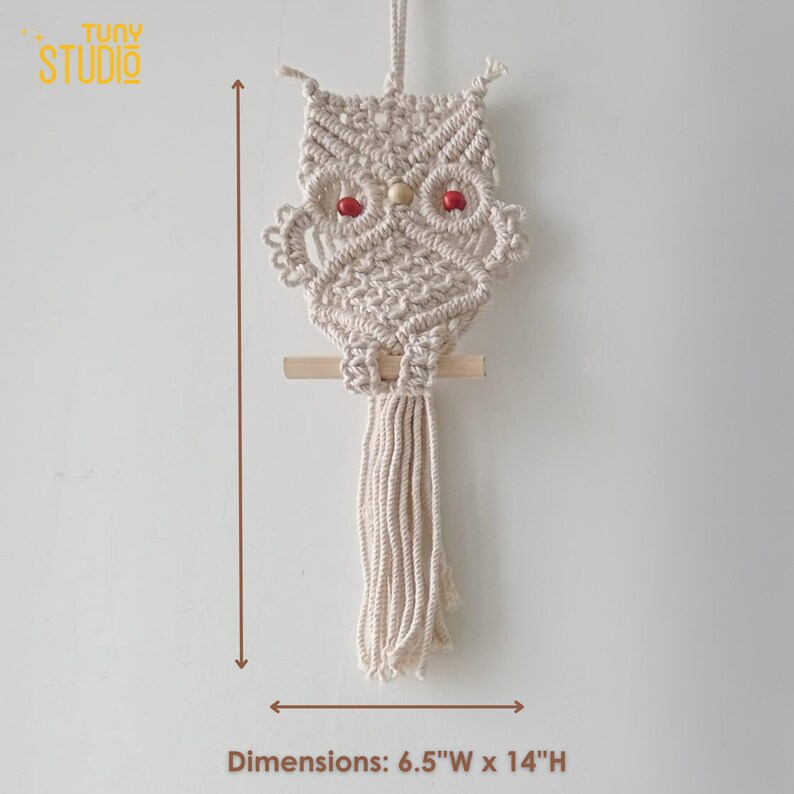 K&ouml;nnte beinhalten: Ein beigefarbener Makramee-Eulen-Wandbehang mit roten Holzperlen als Augen. Die Eule ist 16,5 cm breit und 35,5 cm hoch.