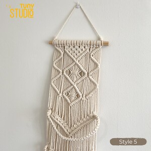 May include: Cream-colored macrame wall hanging with geometric diamond and chevron patterns. The wall decor is suspended from a wooden dowel and features the text "Tuny Studio" in yellow. The style is labeled "Style 5".