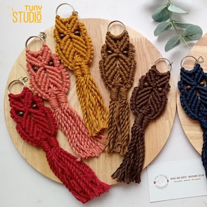 Könnte beinhalten: Sechs bunte, owl-förmige Makramee-Schlüsselanhänger mit Holzperlen für die Augen. Die Schlüsselanhänger werden auf einer Holzoberfläche mit einem kleinen Schild präsentiert, auf dem "Wood and Ropes Macrame Decor" steht.